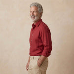 A smiling older man with gray hair and a beard, wearing a red long-sleeve hemp button-up shirt and khaki pants, standing against a textured beige studio background.