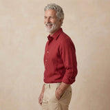 A smiling older man with gray hair and a beard, wearing a red long-sleeve hemp button-up shirt and khaki pants, standing against a textured beige studio background.