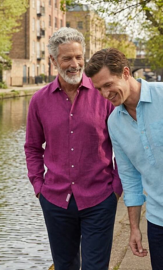 Two men wearing hemp shirts sharing a moment together standing by a river with trees and buildings in the background