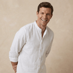 Man wearing a white hemp shirt against a beige background leaning in and smiling at camera whilst adjusting cuffs